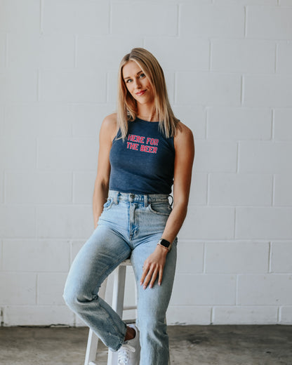 Woman sitting on a stool wearing a tank top with "Here for the Beer" on it and jeans against a white brick wall.